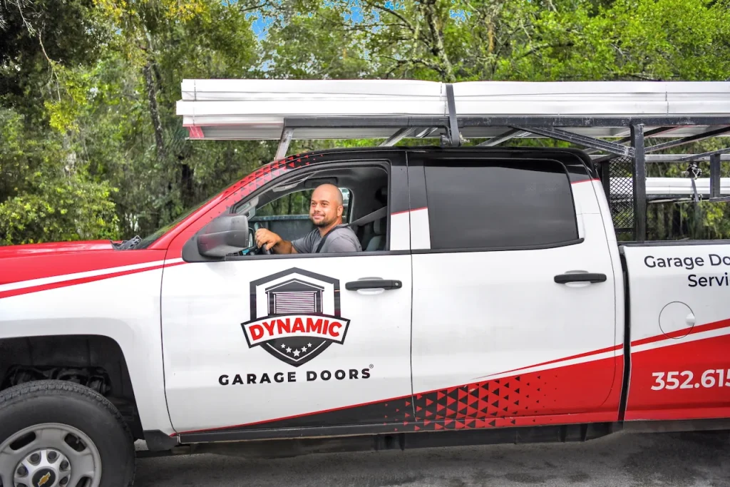 A service technician smiles from a branded company truck equipped with roof racks, ready to arrive for professional garage door maintenance.