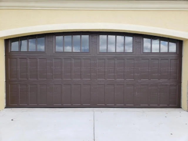 A dark brown arched panel door with upper window grids fits cleanly beneath an exterior stucco archway, showcasing durable garage door installation materials.