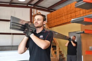 Two technicians carry heavy metal track components inside a workshop, preparing materials for professional garage door services DeLand, FL.