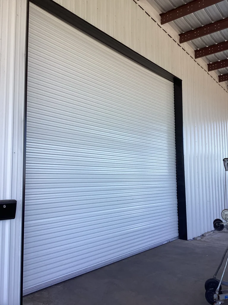 A white corrugated roll-up door with a dark frame is installed on a metal warehouse building, highlighting industrial rolling steel doors.