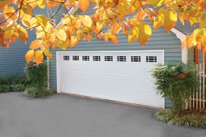 A classic white traditional panel garage door with decorative upper window grids on a blue house, showcasing sleek and secure storm-rated garage door systems.