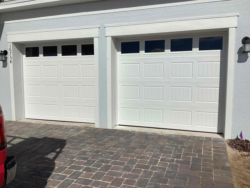 Two white traditional long-panel doors with sleek top window inserts sit side-by-side on a paved driveway, showing an exterior view of a setup requiring heavy-duty garage door springs.