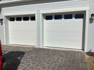 Two white traditional long-panel doors with sleek top window inserts sit side-by-side on a paved driveway, showing an exterior view of a setup requiring heavy-duty garage door springs.